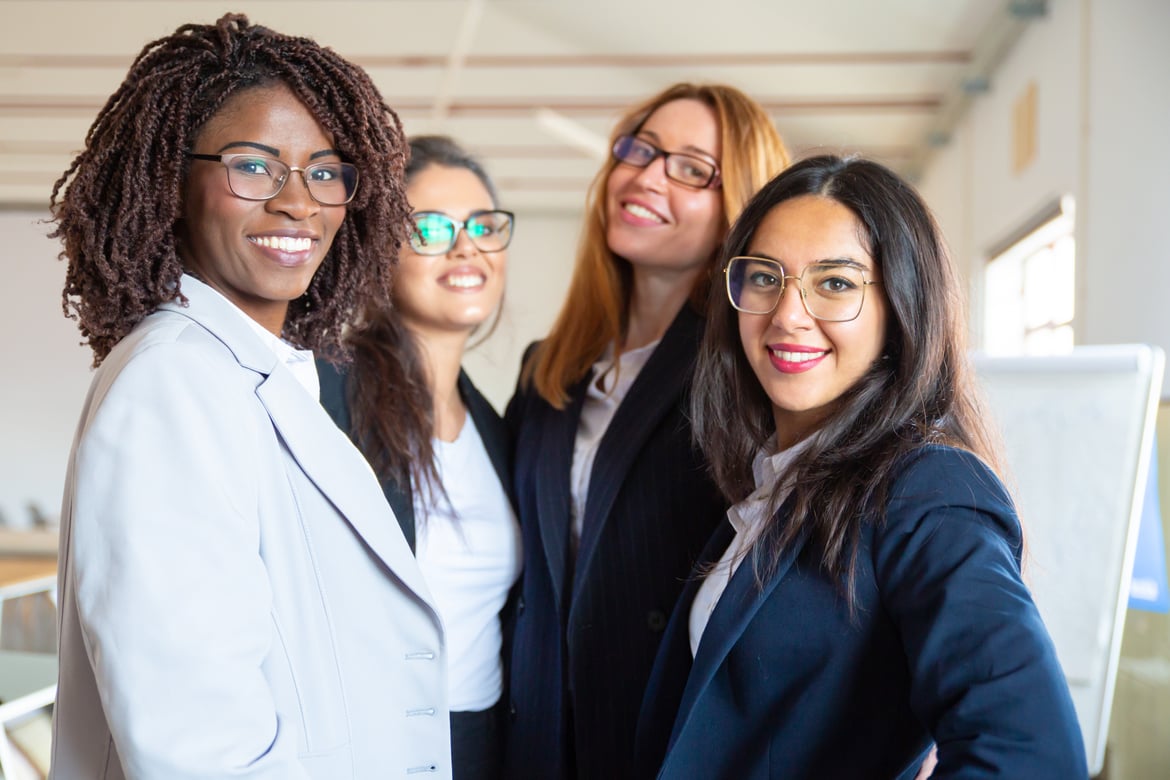 Four confident businesswomen smiling at the camera in a modern office environment.